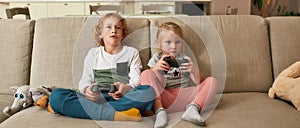 Two siblings, little boy and girl looking focused, playing video games using joystick or controller while sitting