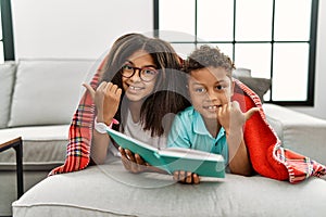 Two siblings lying on the sofa reading a book pointing to the back behind with hand and thumbs up, smiling confident