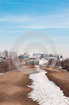 Two silhouettes of people are walking on top of the hill with a line of melting snow leading towards them. A big city is on the