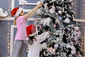 Two sister girls in red caps and pajamas decorate the Christmas tree with balls