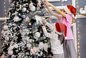 Two sister girls in red caps and pajamas decorate the Christmas tree with balls