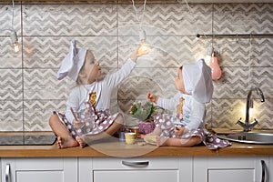 Two sisters in a cook hats sit on the kitchen table and drink tea with cookies