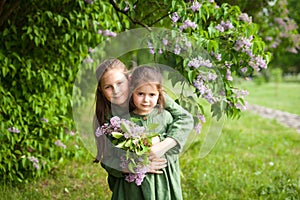Two sisters in green linen dress have fun in the park with blooming lilacs, enjoy spring and warmth
