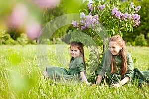 Two sisters in green linen dress have fun in the park with blooming lilacs, enjoy spring and warmth