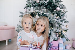 Two sisters at home with Christmas tree and presents. Happy children girls with Christmas gift boxes and decorations.