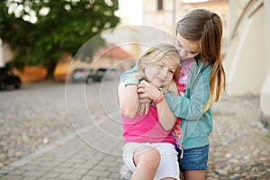 Two sisters laughing and hugging on warm and sunny summer day in a city. Cute siblings having a good time together