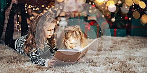 Two sisters are reading holding a book in their hands. Christmas mood. In the background a decorated Christmas tree with Christmas
