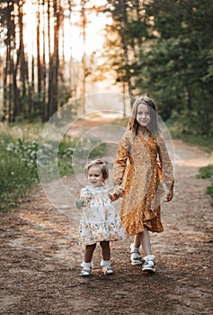 Two sisters run through the forest holding hands. Summer sunny day and girls in light dresses