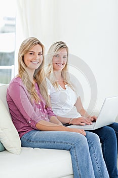 Two sisters sit on the couch with a laptop as they smile at the