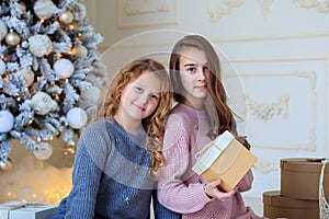 Two sisters sitting near Christmas tree with gift boxes at home.