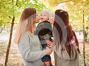 Two sisters of the twins, stand in an autumn park, kiss on both cheeks of a little boy, whom one of them is holding.