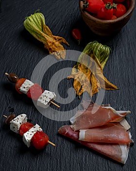 Two skewers with pieces of cheese and cherry tomatoes on a slate plate, with courgette flowers and sliced ham next to them