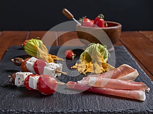 Two skewers with pieces of cheese and cherry tomatoes on a slate plate, with courgette flowers and sliced ham next to them