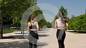 Two slender young women run through a modern park in summer. Sport.