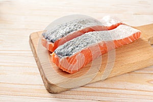 Two slices of fresh salmon fillet with skin on kitchen board and a light wooden table, ingredient for a healthy fish meal, cooking