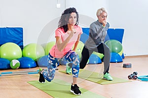 Two slim Caucasian women exercising on mats doing lunges with arms out in front of them in fitness club