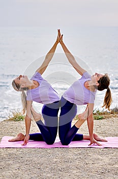 Two slim european sportswomen wearing equal the same activewear doing acro tandem yoga on mat on coast near the sea in the morning