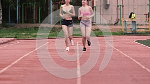 Two slim sporty girls in shorts running around the stadium in slow motion.