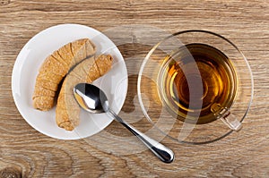 Two small croissants in saucer, spoon, cup of tea on saucer on table. Top view