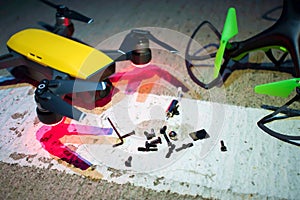 Two small drones lie on the ground. The yellow drone is on and is preparing to take off. Small parts are strewn around two drones