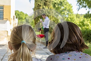 Two small girls are watching a performance of a magician through an outdoor birthday party