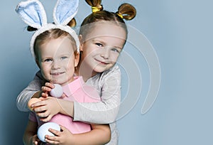 Two small happy girls in cosy home clothing and decorative fur ears standing, hugging and holding white and beige eggs