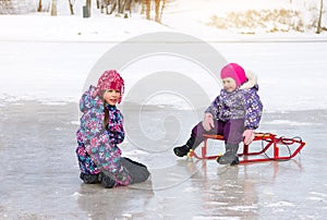 Two small kids have fun sitting together on the ice and playing with a snow sled on clear winter day