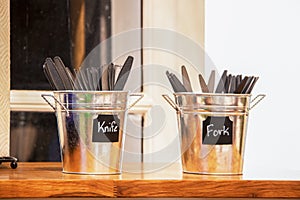 Two small tin buckets on counter at restaurant holding plastic cutlery - knives and forks - selective focus with bold blocked