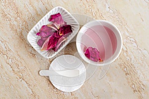 Two small white bowl with rose water toner, dry rose petals and cotton pad. Ingredients for preparing homemade cosmetics.