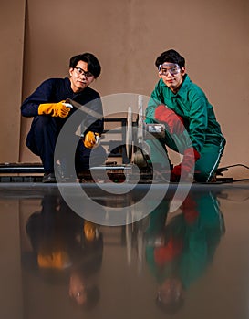 Two smart Asian engineers wearing a mechanic jumpsuit, safety glasses, and glove sitting and holding tools posing smiling to a