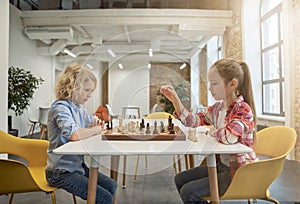 Two smart little children, boy and girl developing chess strategy, playing board game, sitting together at the table in
