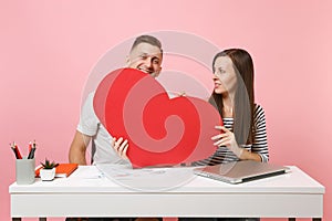 Two smiling business woman man colleagues sit work at white desk with contemporary laptop on pastel pink background.