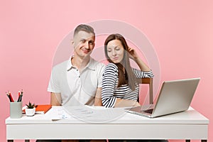 Two smiling business woman man colleagues sit work at white desk with contemporary laptop on pastel pink background.