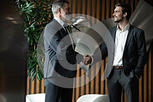 Two smiling businessmen shaking hands together while standing by windows in an office boardroom.