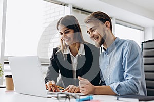 Two smiling businesspeople sitting together at a table in a modern office talking and using a laptop