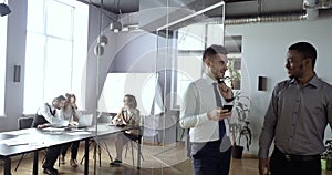 Two smiling colleagues of man stand in corridor looking at funny video on phone, chatting laughing, having fun at work