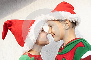 two smiling cute children in Santa hats in profile looking at each other, on a light background