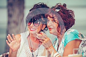 Two smiling female senior friends talking and pointing fingers to someone while drinking coffee