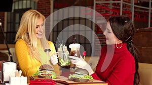 Two smiling girls drink cocktails and eat burgers in a bright restaurant.