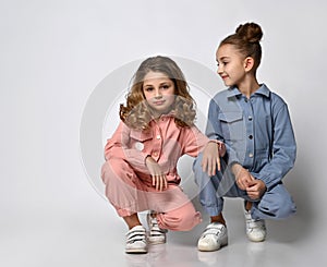 Two smiling girls, friends, sisters in blue and pink cotton overalls with pockets are squatting posing for the camera.