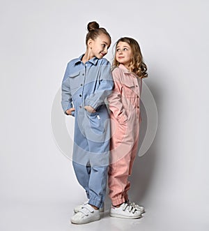 Two smiling girls, friends, sisters in blue and pink cotton overalls with pockets stand close to each other, looking at