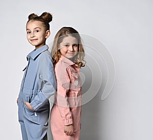 Two smiling girls, friends, sisters in blue and pink cotton overalls with pockets stand close to each other, looking at