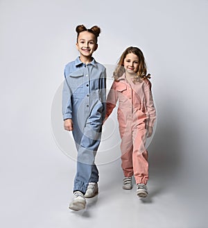 Two smiling girls, friends, sisters in blue and pink cotton overalls with pockets walk towards us on a light background