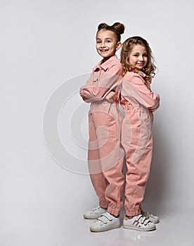 Two smiling girls, friends, sisters in pink cotton overalls with pockets stand close to each other, looking at the