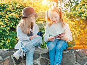 Two smiling little sister girls kids sitting and browsing their smartphone devices in the autumnal park. Careless young childhood