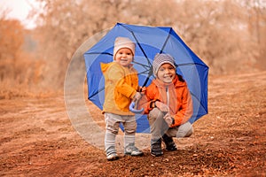 Two smiling little toddlers, brother and sister, hiding together under big blue umbrella in autumn park