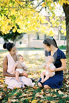 Two smiling mothers with babies on their laps sit on yellow foliage in the park
