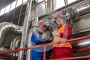 Two Smiling Oil Refinery Workers in Protective Workwear with Thumbs Up Beside Refinery Piping