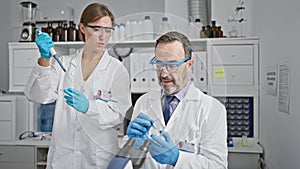 Two smiling scientists, mates in science, pouring liquid into a test tube using a pipette in an indoor lab, zoom in on their