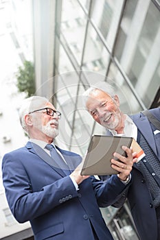 Two smiling senior businessmen working on a tablet standing in front of an office building